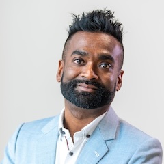 A suave dapper man with brown eyes, a close-cropped dark beard and punky hair in a stylish light blue suit against a white background.