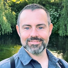 A bearded short-haired man with light skin smiling outdoors amongst greenery.