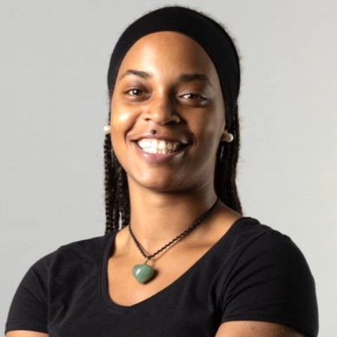 A smiling dark-skinned young woman with long hair wearing a black T-shirt and a green pendant in front of a light background.