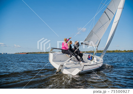 A beautiful white yacht is sailing in the wind on the river against the background of a beautiful autumn forest. A guy on board with two girls. 75500927