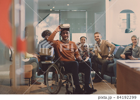 Disabled businessman in a wheelchair at work in modern open space coworking office with team using virtual reality googles drone assistance simulation 84192299