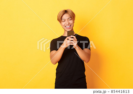 Portrait of handsome stylish asian guy with blond hair, using mobile phone and smiling, messaging in smartphone app, yellow background 85441613