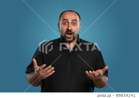 Close-up portrait of a brunet middle-aged man with beard, dressed in a black t-shirt and posing against a blue background. 89834241