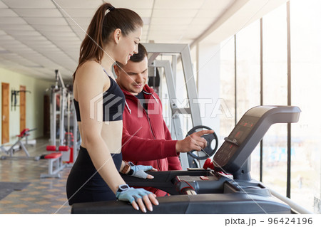 Cheerful magnetic young lady asking for help her trainer, trying to use running machine, listening to instructions carefully. Attractive girl wears black tight top, shorts and light blue gloves. 96424196