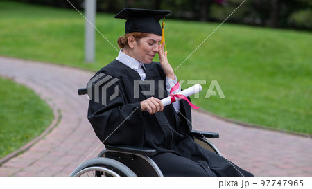 A caucasian woman in a wheelchair holds her diploma and cries with joy outdoors. 97747965