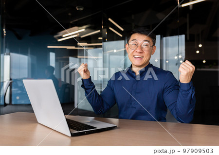 Successful asian businessman celebrating victory success, portrait of man in office in casual shirt, employee smiling and looking at camera satisfied with achievement holding hand up victory gesture. 97909503