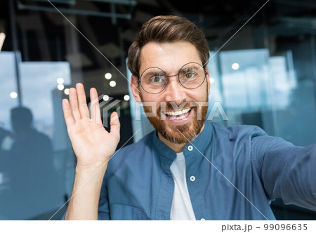 Mature man in office wearing shirt and glasses smiling and looking at smartphone camera, businessman taking selfie and talking on video call using smartphone, programmer waving hand greeting gesture. 99096635