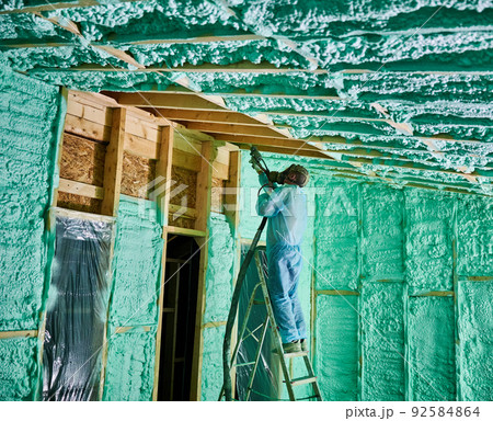 Male builder insulating wooden frame house. Man worker spraying polyurethane foam inside of future cottage, standing on ladder, using plural component gun. Construction and insulation concept. 92584864