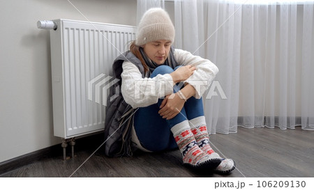 Young woman wearing lots of warm clothes sitting on floor next to heater in cold apartment 106209130