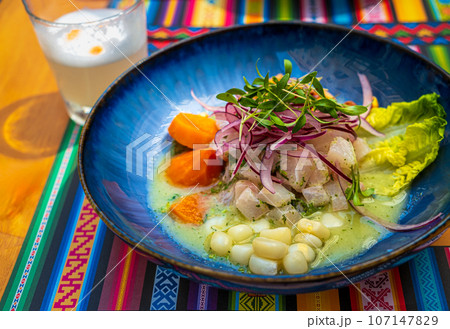 Fresh ceviche served in blue plate over placemat with traditional patterns and Pisco Sour cocktail. Selective focus, blurred background. Peruvian-style ceviche made out of raw fish, red onions, corn 107147829