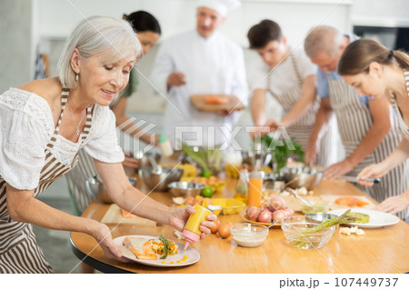 Elderly woman seasoning dish with sauce during group cooking class 107449737