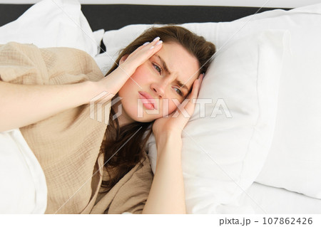 Close up portrait of sick woman lying in her bed and feeling unwell, touching head, has headache, looking frustrated from migraine, wearing pyjamas 107862426