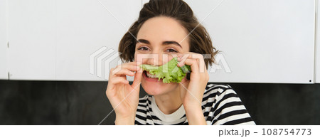 Close up portrait of young woman, vegetarian girl, likes eating vegetables, posing with lettuce leaf and smiling, posing in the kitchen. Concept of healthy food and diet 108754773