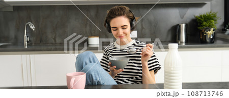 Portrait of smiling young candid woman, student eating morning cereals, having her breakfast, listening music in wireless headphones, sitting in the kitchen 108713746