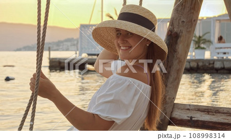 Portrait of happy smiling woman in straw hat sitting on the swing against sunset over the sea beach 108998314