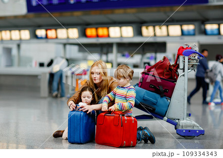 Two tired little kids, boy and girl, siblings and mother at the airport. Children, family traveling, going on vacation by plane and waiting by luggage trolley with suitcases at terminal for flight. 109334343