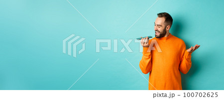 Happy man talking on speakerphone, gesturing and recording voice message on mobile phone, standing in orange sweater over light blue background 100702625