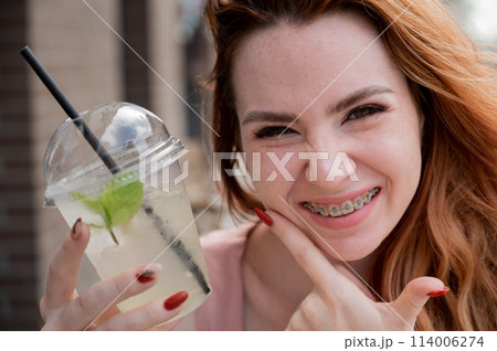 Young beautiful red-haired woman with braces drinks cooling lemonade outdoors in summer. Portrait of a smiling girl with freckles. 114006274