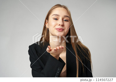 Young Woman in Business Attire Gesturing a Kiss With Hand Against a Gray Background 114507227