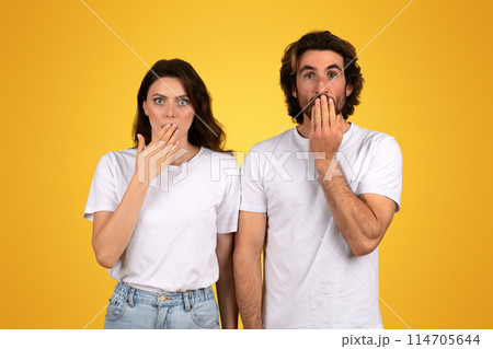 Shocked millennial european man and a woman with surprised expressions, both covering their mouths with their hands, dressed in white shirts against a yellow background, studio 114705644