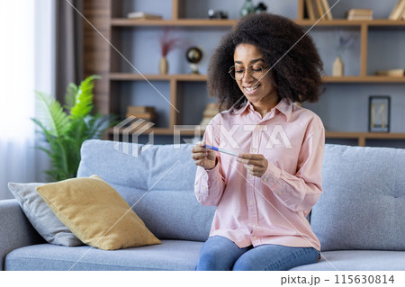 Happy woman holding a positive pregnancy test while sitting on a couch in a comfortable and cozy living room. 115630814