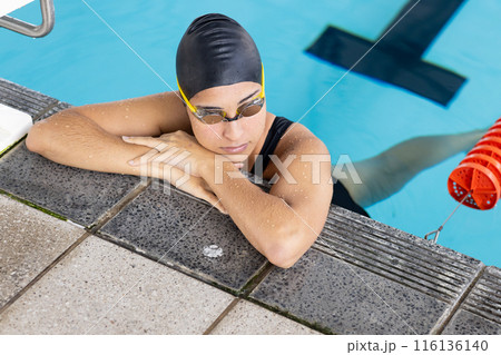 Biracial young female swimmer resting at pool edge indoors, wearing goggles 116136140