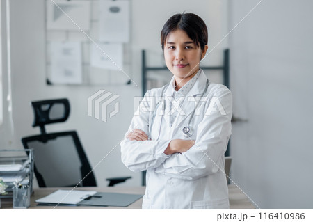 A confident female doctor in a white coat with a stethoscope stands in a modern medical office, ready to assist patients. 116410986