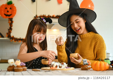 Happy mother and daughter dressed in Halloween costumes decorating cupcakes together in decorated kitchen 116420117