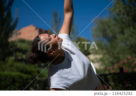 Woman practicing yoga outdoors, stretching arm under blue sky in serene natural setting 116425282