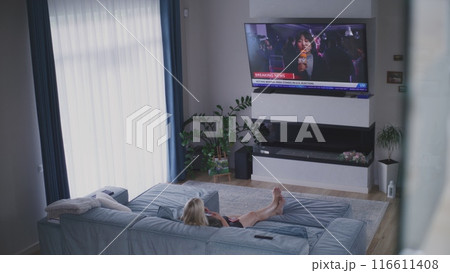 Woman rests in living room, watches live broadcast on TV 116611408