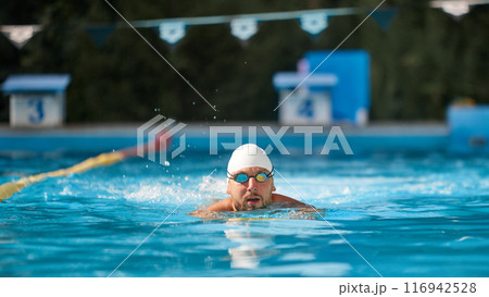 Determined swimmer in white cap and goggles glides through pool, focused on his training against blurred sunny outdoor background. 116942528