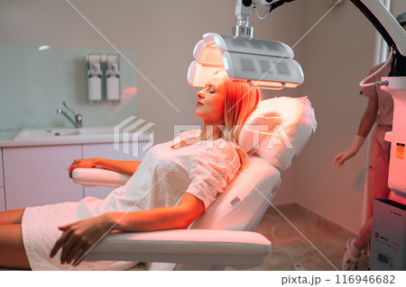 Side view of woman client with blonde hair lying chair having LED light hair PDT treatment at luxury aesthetic clinic. Female specialist adjusting innovation device for photodynamic therapy procedure 116946682