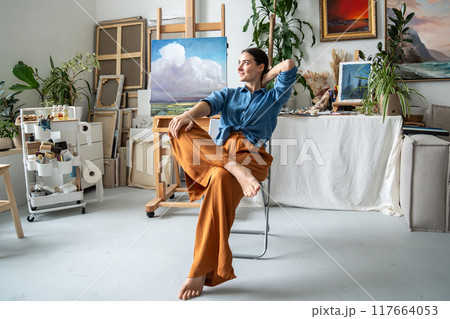 Dreaming pleased woman artist takes break from creative painting process sits on chair in art studio 117664053