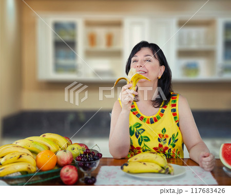 Middle-Aged Woman in Colorful Outfit Eating Banana in Kitchen, Showcasing the Joy and Health Benefits of a Fruit-Rich Diet. 117683426