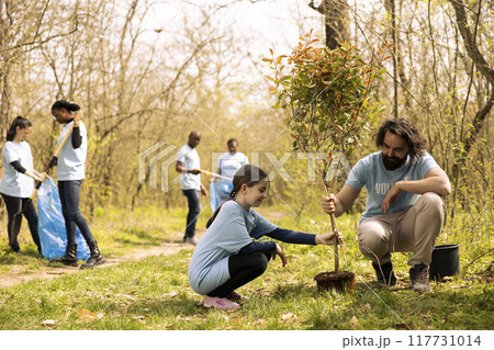 Man and young kid volunteering to fill a hole with a tree, working on reforestation mission together. Proud father and daughter planting greenery to increase forest vegetation. 117731014