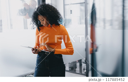 Intently focused Black businesswoman in an orange sweater navigating a tablet within a modern 117885452