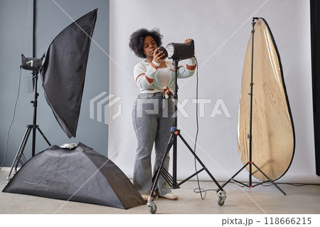 Full length portrait of young creative African American woman as female photographer setting up lighting equipment in studio copy space 118666215