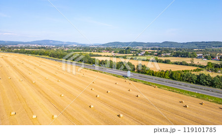 Aerial perspective showing a field of hay bales beside a highway, surrounded by green trees and distant hills under a clear blue sky. 119101785