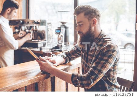 Working at coffee shop. Side view of young handsome man using his digital tablet while sitting at bar counter at cafe with barista at background 119128751