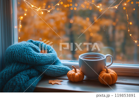 A cup of hot tea wrapped in soft knitted scarf near the window with autumn landscape. Light garland and pumpkins, warm and cozy autumn atmosphere, hello, autumn theme 119853891