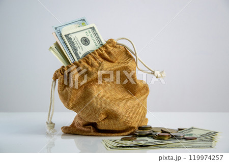 A burlap sack filled with US dollar bills and coins rests on a white surface, with a few bills spilling out 119974257