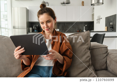 Portrait of smiling beautiful woman, sitting on couch, watching videos on digital tablet, reading e-book on her device, resting at home 110760899