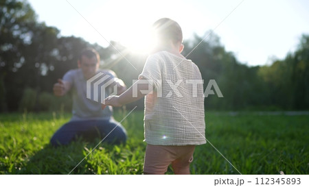 baby takes first steps in the park. happy family kid dream concept. dad teaches his son to walk takes the first steps lifestyle in the park on green grass. baby son taking first steps 112345893