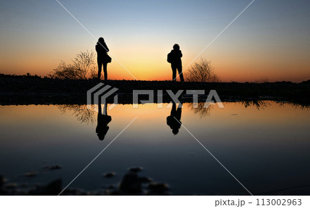 Reflections in the water of silhouette female photographers taking sunrise photos 113002963