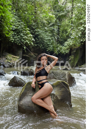 A young slender woman in a swimsuit poses on a huge boulder covered with moss in the middle of a mountain river in the jungle on the popular island of Bali. 120824997