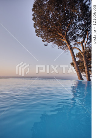 An infinity edge pool with trees against a blue sky background with copyspace. Beautiful outdoor swimming pool at a resort or luxury holiday villa against. Romantic getaway with a sea view in summer 121056058