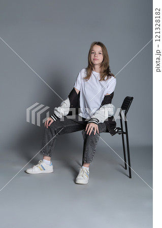 A teenage girl sits casually in a chair, her gaze steady and assured. Her posture reflects the self-confidence and ease of todays youth, embracing a minimalist style. 121328182