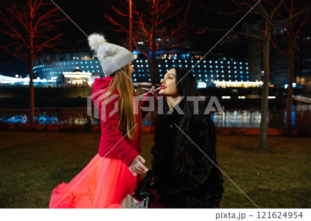 Mother and daughter enjoying a night out at the park with festive lights in the background during winter 121624954