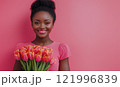 Smiling woman holds vibrant tulips against a pink backdrop during a bright, sunny day 121996839