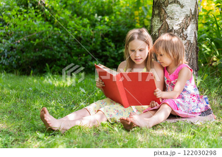 Two sisters reading a red book together in a sunny garden setting 122030298